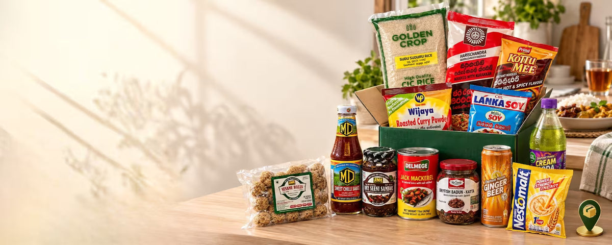 A curated food gift box and assortment of packaged snacks, sauces, rice, and beverages arranged on a wooden table in a warm indoor setting. Visible items include Golden Crop rice, Vijaya boosted curry powder, Kotu Mee spicy pap, Lanka Soy, MD hot sauce, Delmeji sambar, Ginger Beer, Nescafe Cream, and other packaged foods in a green box.